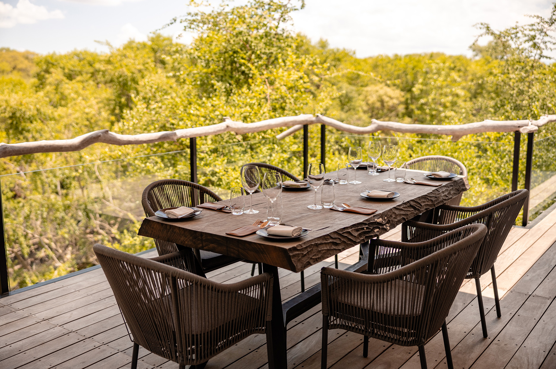 Outdoor dining setup on a wooden deck overlooking the lush treetops at Kapama, featuring a rustic table with place settings and wine glasses.