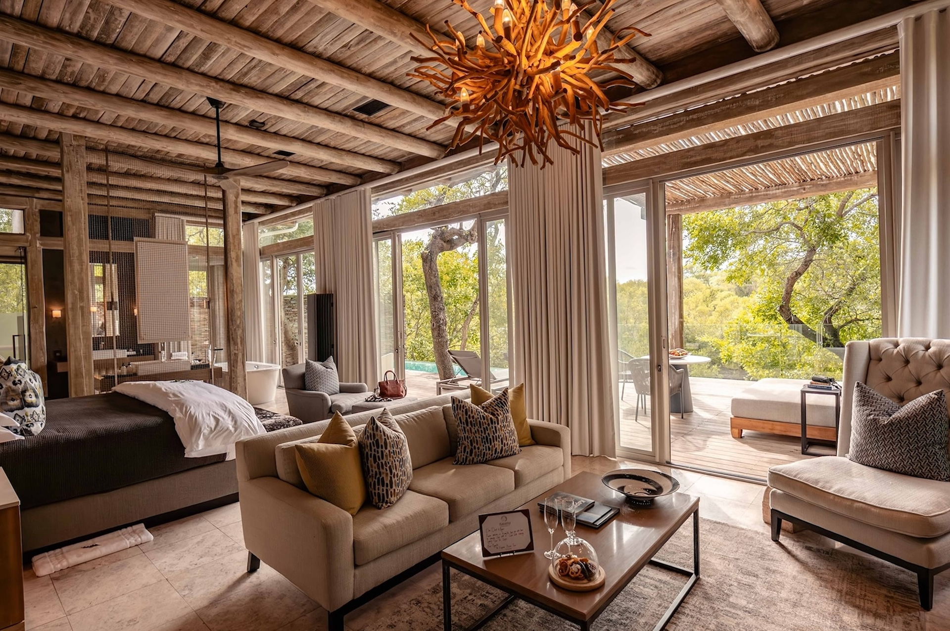 Elegant safari suite at Kapama with open-plan bedroom and lounge, modern rustic decor, forest views, and private deck access through sliding glass doors.