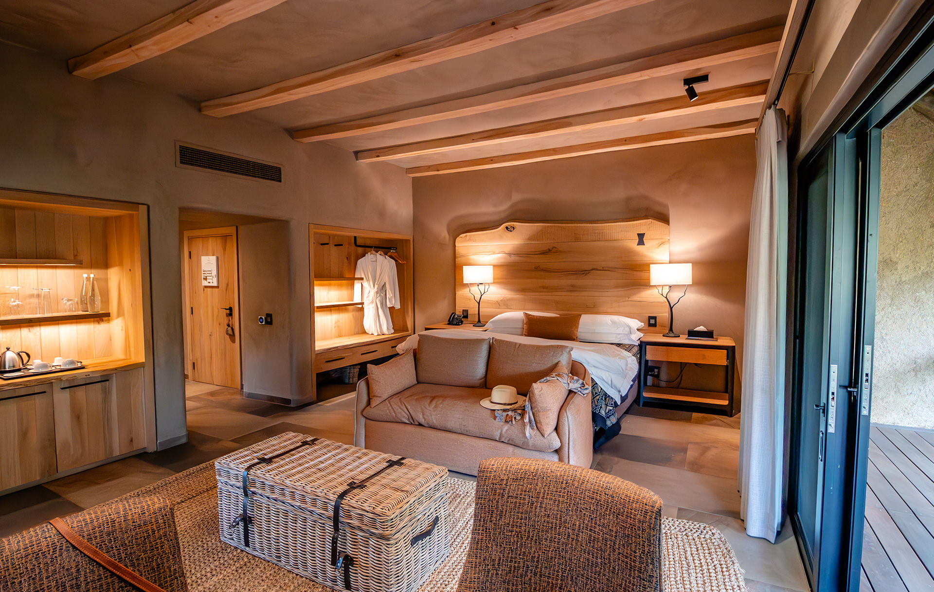 Stylish safari suite with natural wood accents, cozy seating area, king-size bed, open wardrobe with robe, and sliding door leading to a private deck.