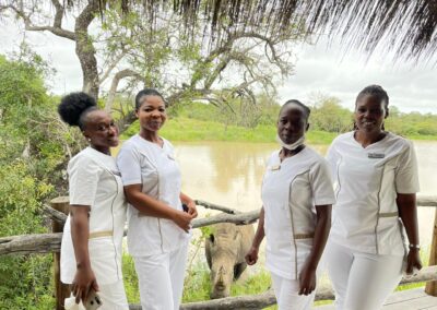 Four Kapama spa team members dressed in white uniforms standing in front of a wooden deck with a rhinoceros drinking from the waterhole behind them.