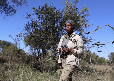 A female safari guide standing in the bushveld with binoculars in hand, dressed in khaki and beige uniform, under a clear blue sky at Kapama Private Game Reserve.