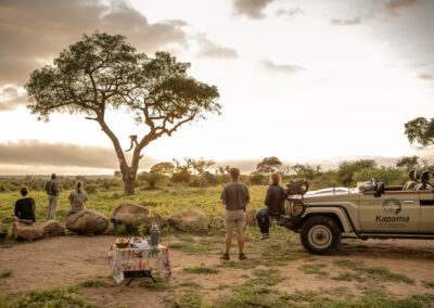 uests enjoying a serene sundowner break in the African bushveld with a Kapama Private Game Reserve safari vehicle parked nearby, overlooking the sunset and savanna landscape.